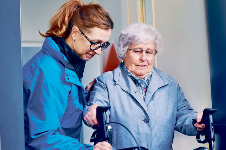 Mitarbeiterin von SPITEX BASEL hilft betagter Frau mit Rollator aus dem Haus 